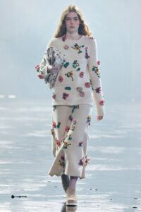 Model wearing a cream knit sweater and matching skirt with colorful 3D floral appliqués.