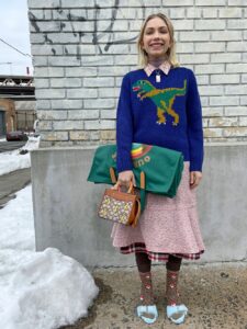 Woman wearing a blue sweater with green dinosaur, pink skirt, and playful winter accessories.
