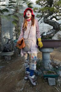 Woman in colorful streetwear with patchwork pants, red hat, and eclectic accessories.