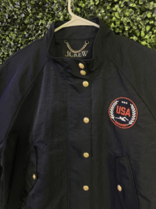 Navy blue J.Crew jacket featuring a circular USA patch on the chest, hanging on a white hanger against a green leafy backdrop.