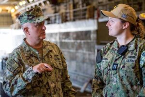 Military personnel and a civilian in conversation in an industrial setting.
