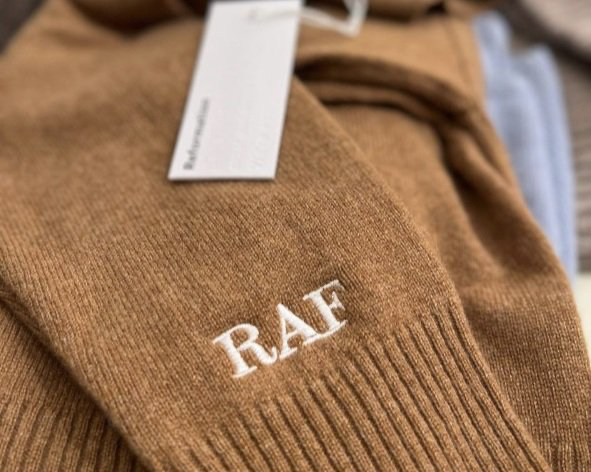 Close-up of “RAF” monogram embroidered in white on a camel-colored knit sweater.