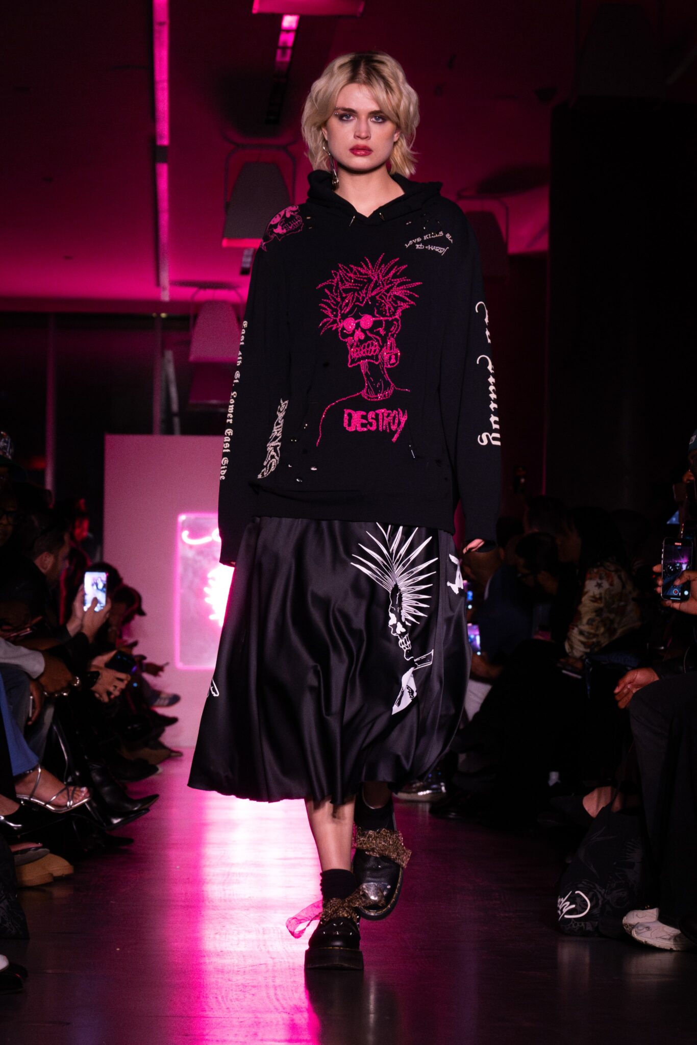 Model wearing black hoodie and skirt featuring bold pink embroidery on a runway.