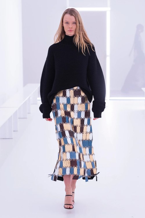 Model in Brandon Maxwell Spring 2024 RTW wearing a loose black turtleneck and colorful rectangular patchwork midi skirt with strappy heels, walking on a bright white runway.