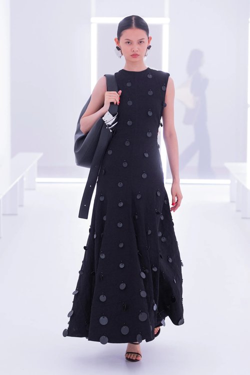 Model in Brandon Maxwell Spring 2024 RTW wearing a sleeveless black dress with tonal dot appliqués, black shoulder bag, and strappy heels on a white runway.