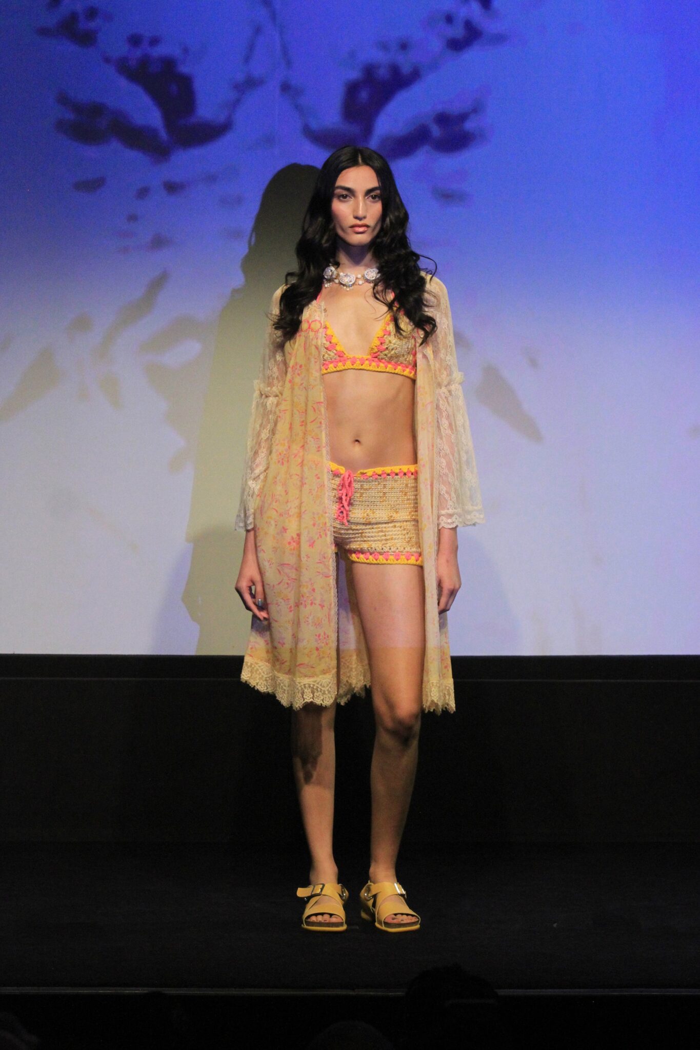 Model wearing a sheer yellow lace robe over orange lingerie on a dark runway.