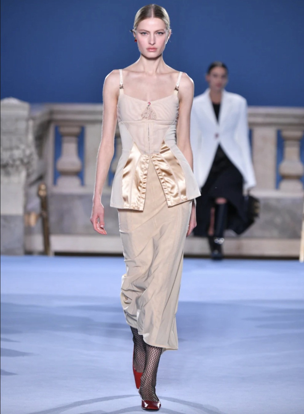 Model walks runway in a nude-toned corset top with ruched satin detail and midi skirt.