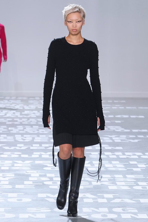 Model in a fitted black long-sleeve knit dress with subtle side drawstrings, styled with black knee-high boots on a light-toned runway.