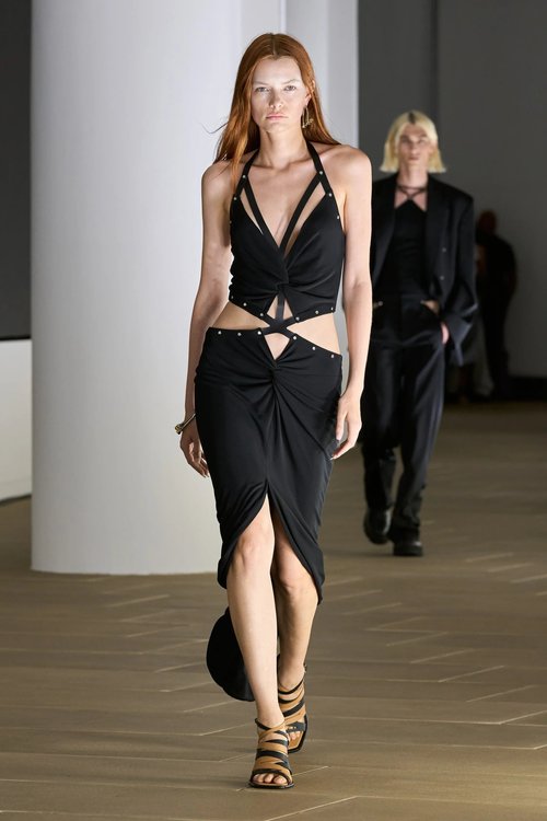 Model wearing a black cutout halter dress with midriff exposure and pointed draping at the skirt, walking in heels.