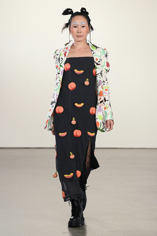 Model in black dress with fruit appliqués and a printed jacket, walking the runway.