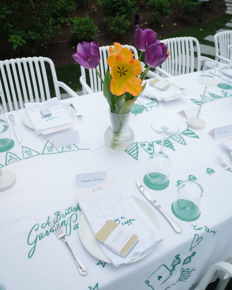 Elegant outdoor table setup with colorful tulips in a clear vase, white linens embroidered with green designs, and neatly arranged place settings for a garden-themed event.