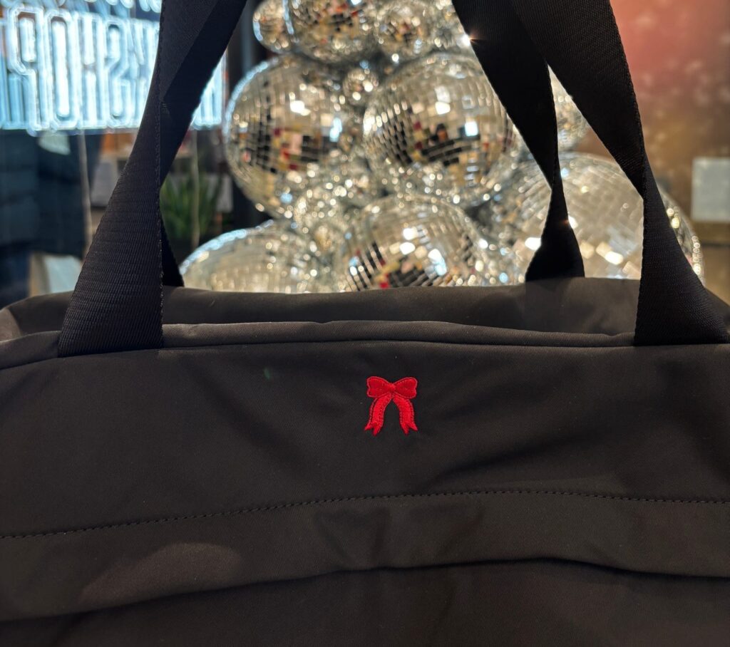 Close-up of a black AWAY tote bag featuring a red embroidered holiday bow, stitched by NYES in front of the disco-ball installation at the Winter Workshop.