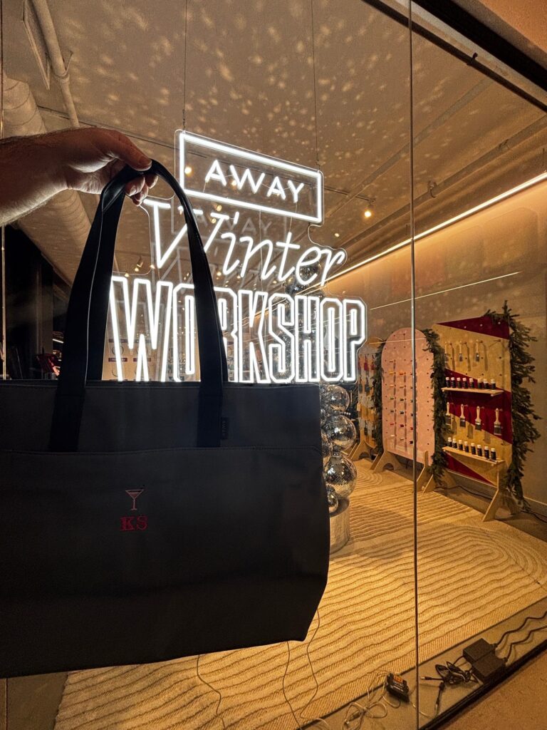 Black AWAY tote bag customized with red embroidered initials “KS” and a martini glass motif, held in front of the illuminated AWAY Winter Workshop sign at the holiday activation.