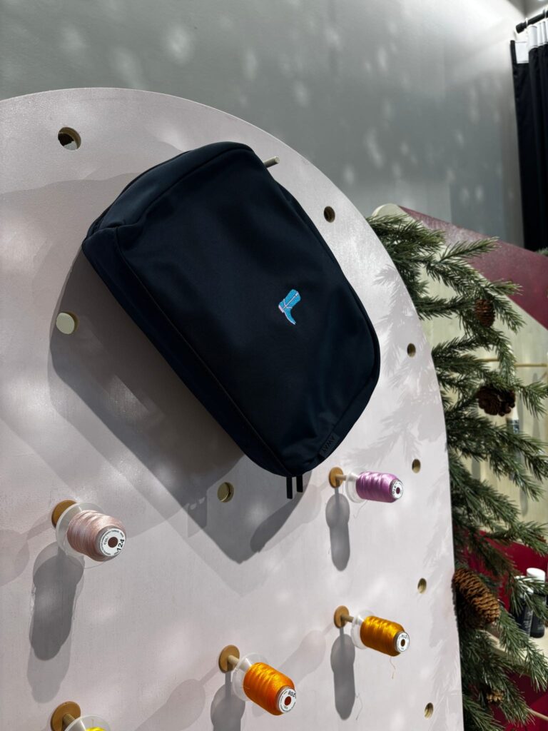 Black AWAY travel pouch customized with blue embroidered motif, displayed on a thread wall created by New York Embroidery Studio during AWAY’s Winter Workshop holiday event.
