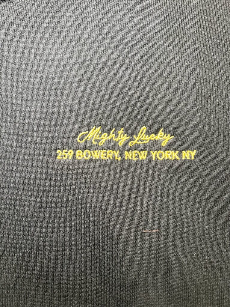 Black hoodie embroidered with Mighty Lucky logo and New York address on the chest