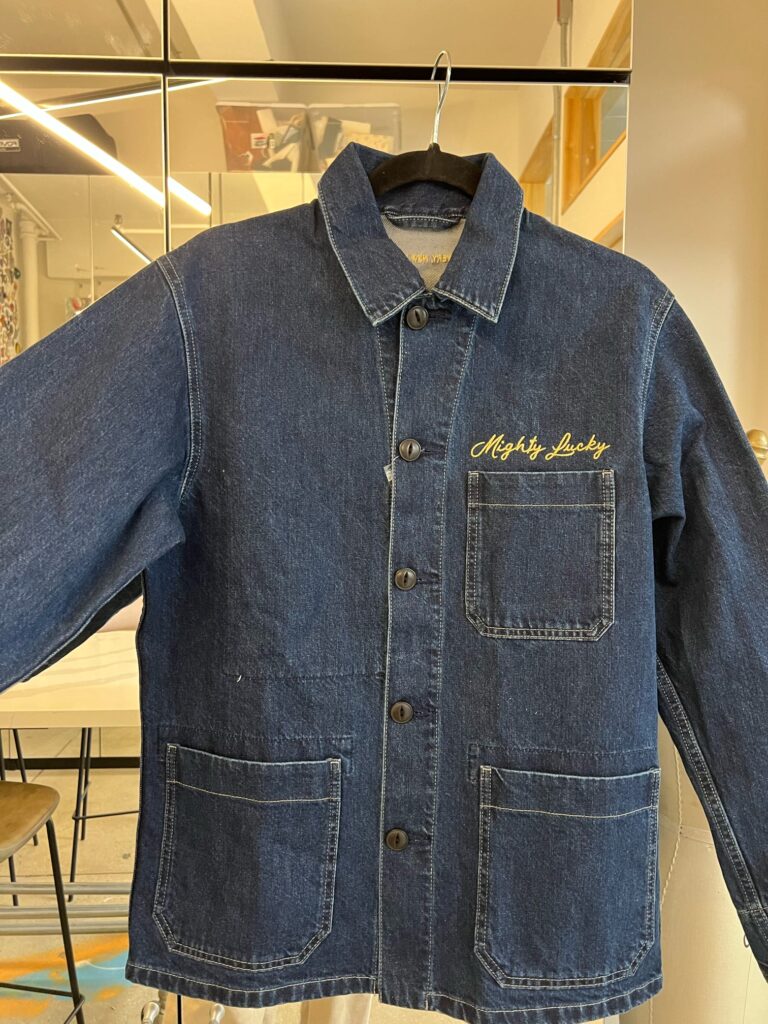Front view of a denim jacket with Mighty Lucky chest embroidery displayed on a hanger