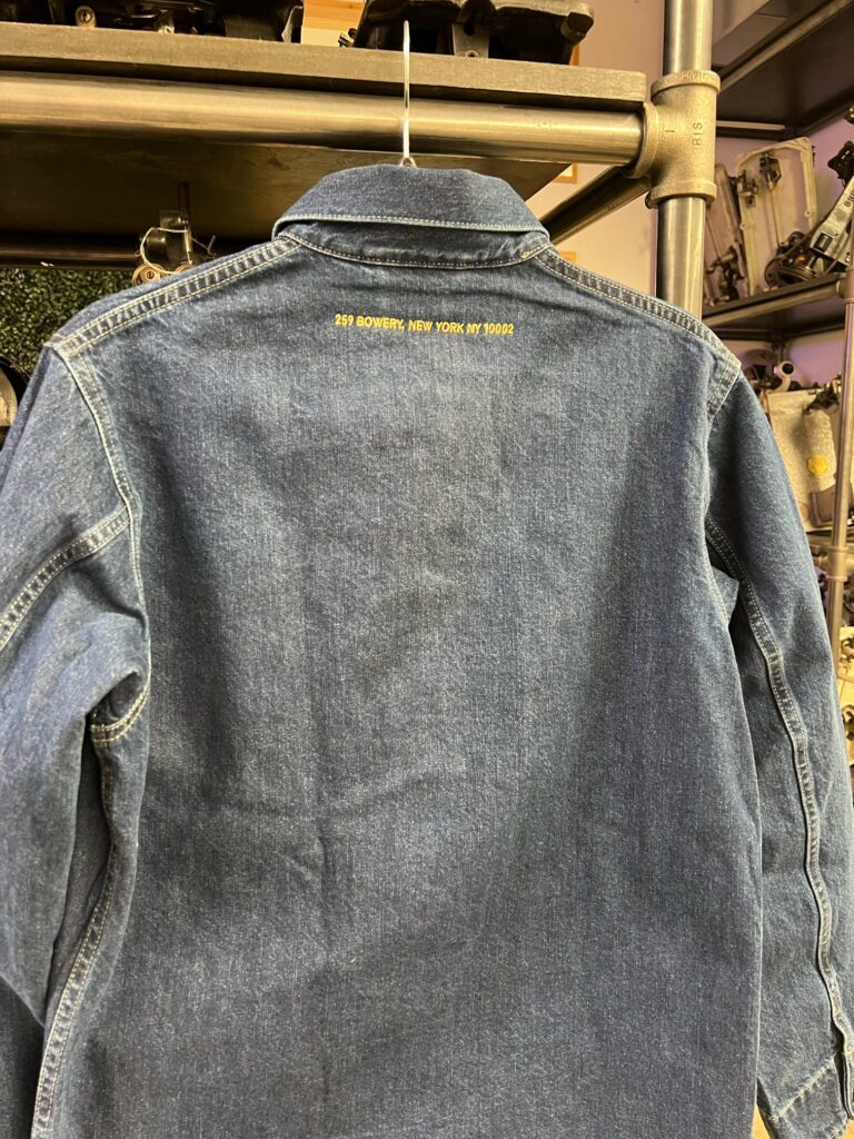 Back view of a denim jacket embroidered with Mighty Lucky and New York address detail in gold thread