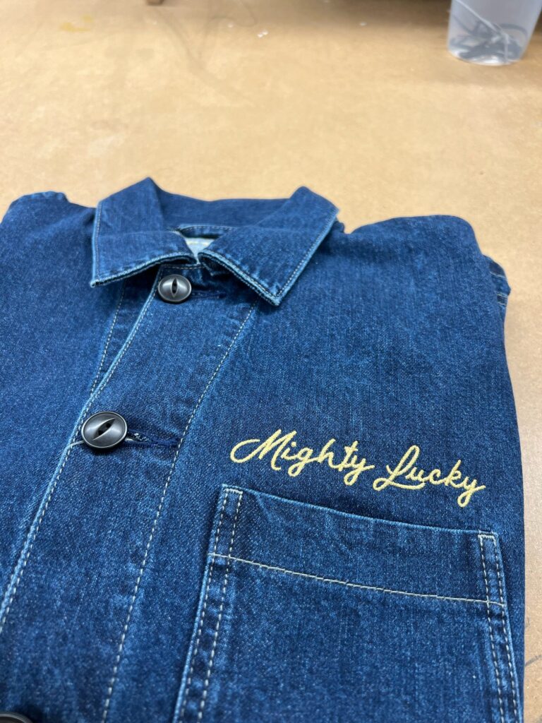 Close-up of Mighty Lucky script embroidery stitched on the chest of a denim jacket