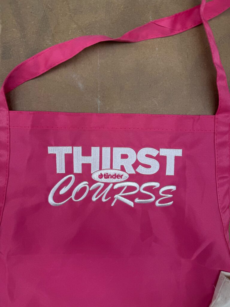 Pink apron embroidered with Thirst Course and Tinder logo in white thread