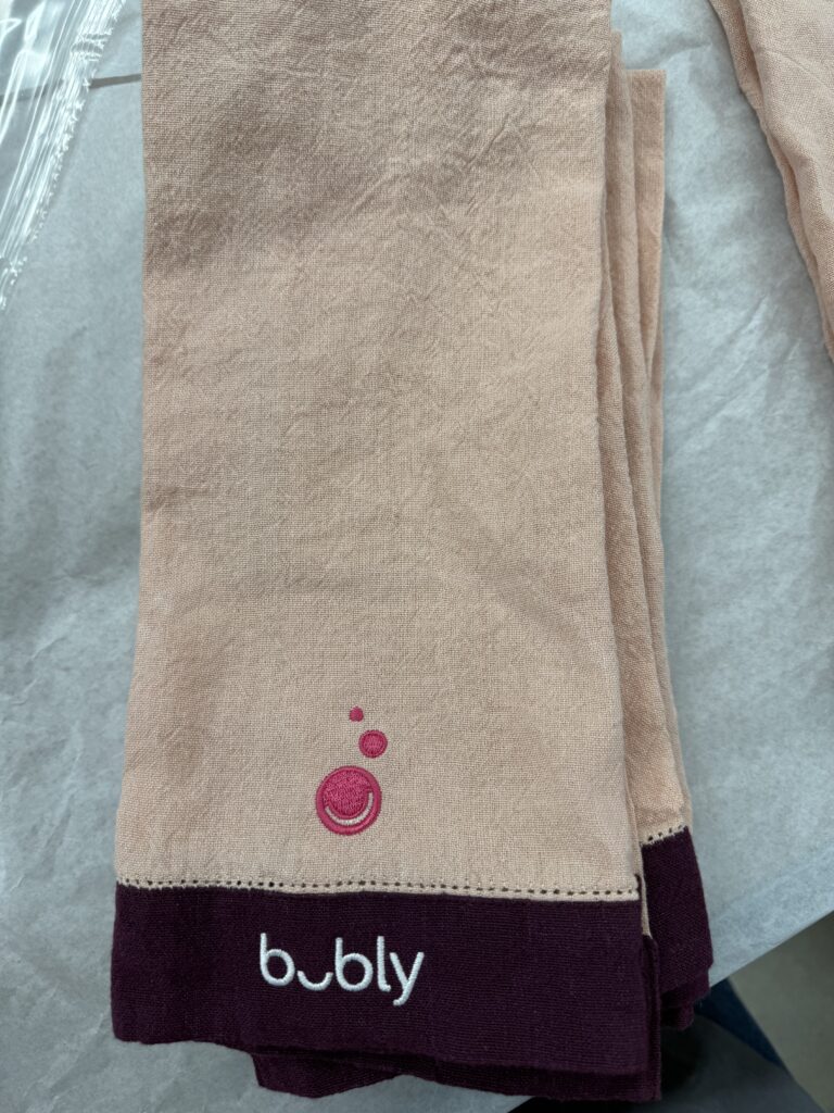 Beige bar towel with burgundy trim featuring custom embroidered bubly logo and icon