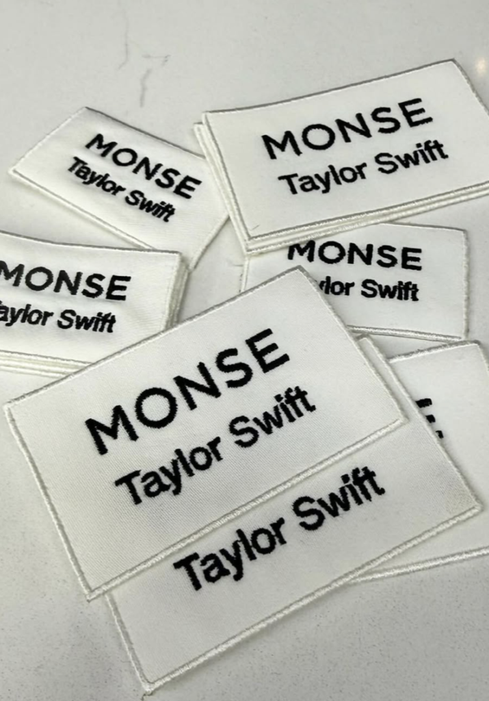 A pile of several scattered white embroidered clothing tags with black text that reads Monse Taylor Swift on a clean white countertop.